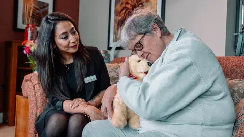 Care home manager comforting a dementia resident with a robotic therapy dog in a lounge
